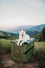 White dog sitting on top of their od green collapsible dog crate in the mountains, with a view of more mountains and trees in the background